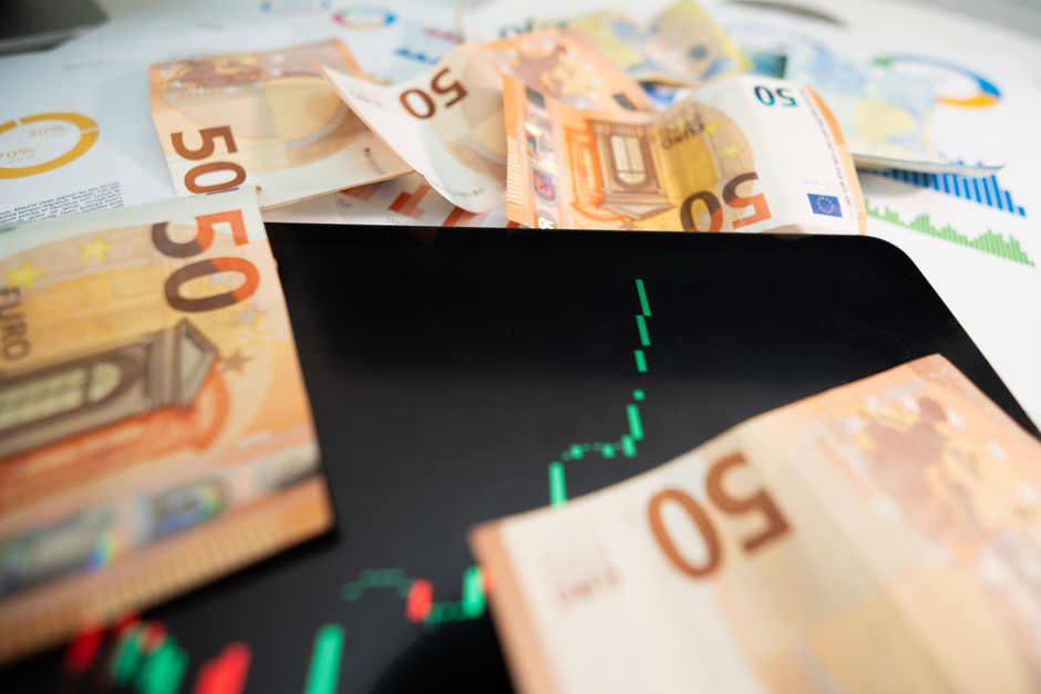 A digital tablet displaying a green financial chart or graph, partially covered by scattered fifty-euro banknotes. The banknotes are crumpled and placed around the tablet, with some overlapping each o